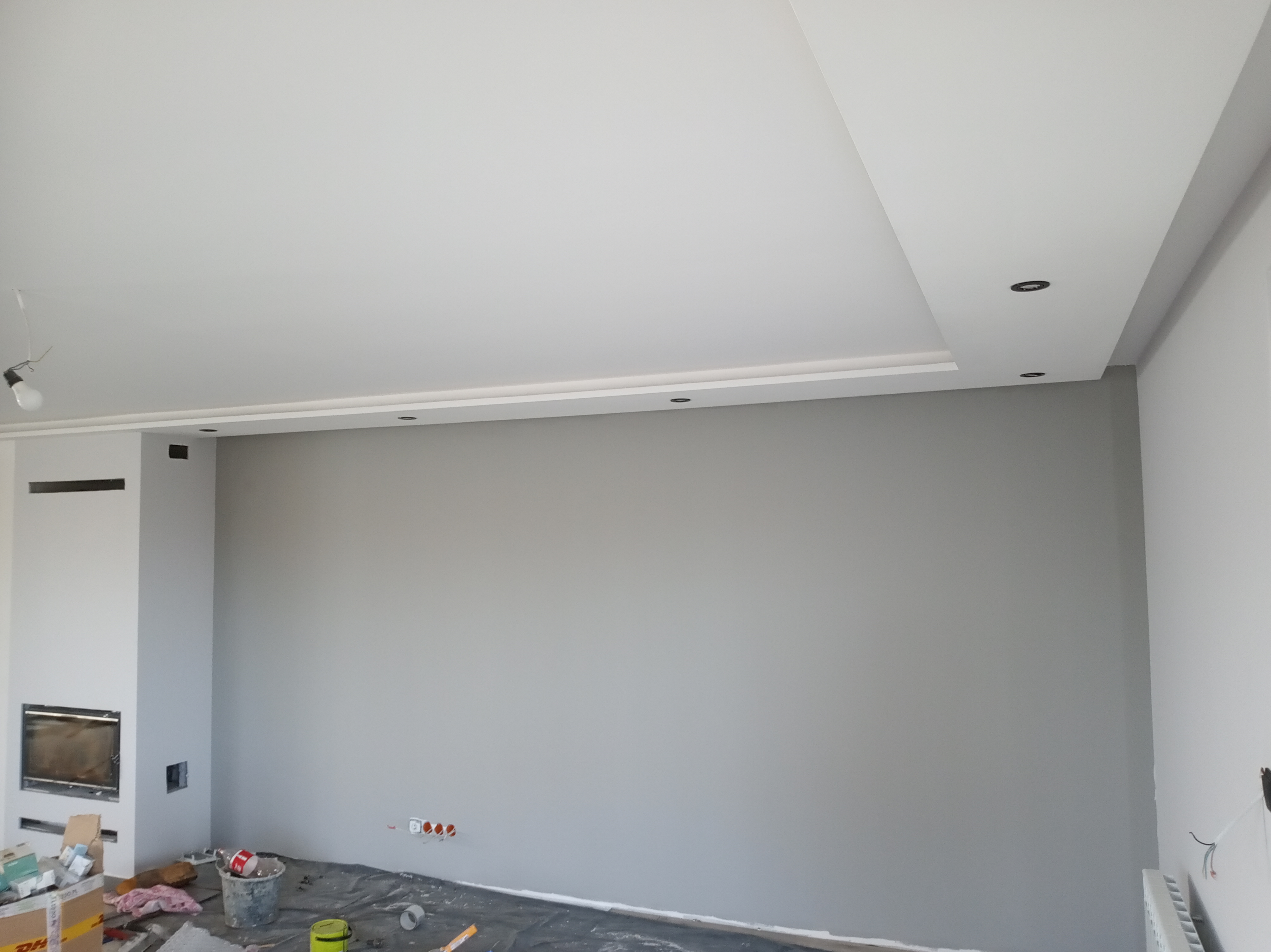 Wnętrze pokoju w trakcie malowania na szaro, z widocznym kominkiem, fragmentem sufitu z oświetleniem punktowym i zabezpieczoną folią podłogą, puszki z farbą oraz narzędzia malarskie w nieładzie.