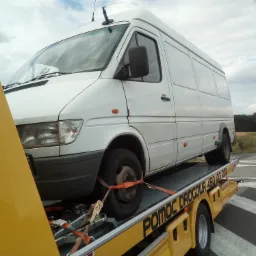 Biały bus transportowany na żółtej lawecie, zabezpieczony pomarańczowymi pasami transportowymi, widoczne oznaczenia na lawecie 'POMOC DROGOWA 450 103 203'.