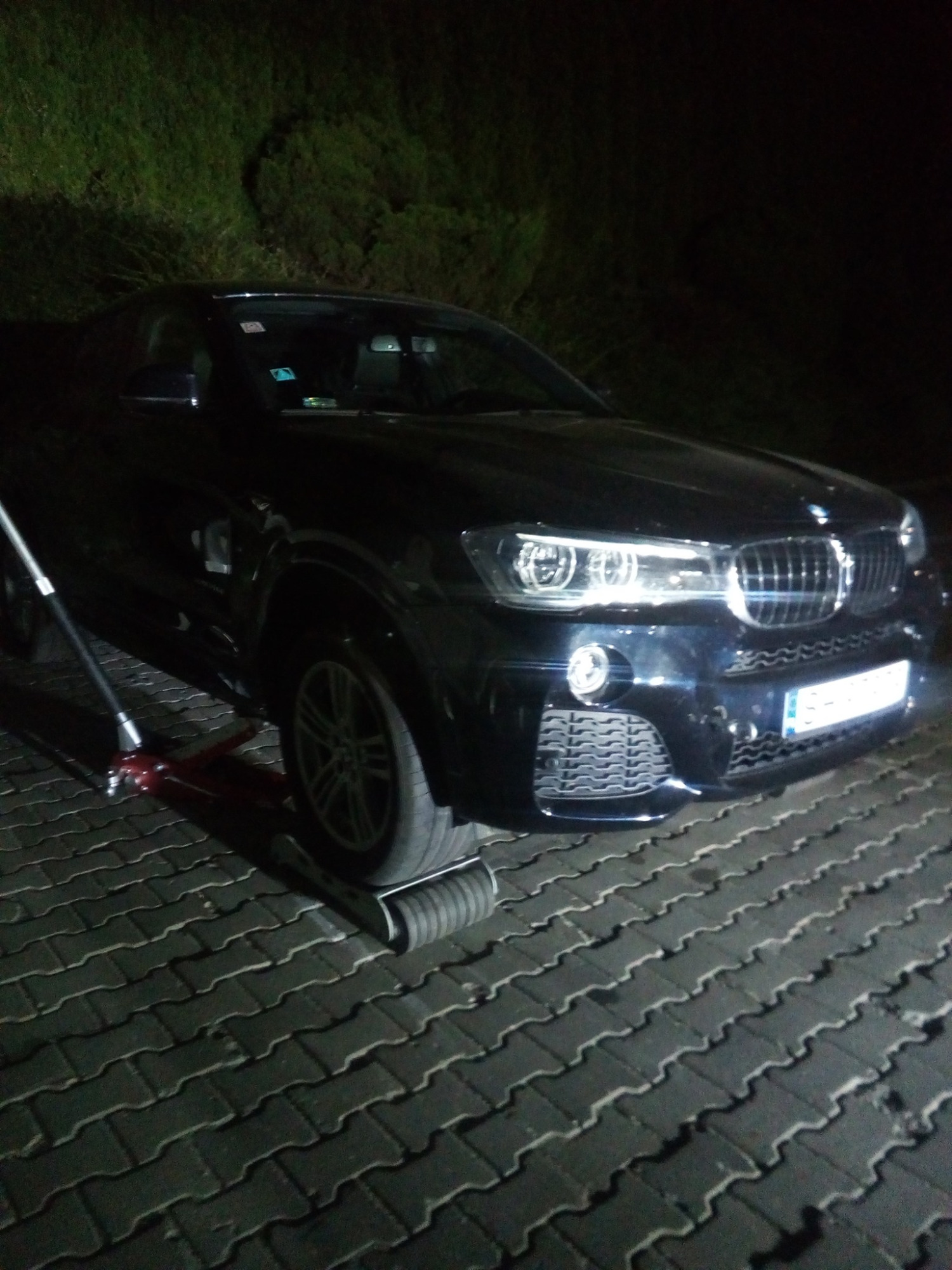 Ciemny granatowy samochód BMW podnoszony za pomocą podnośnika hydraulicznego na brukowanej powierzchni, z widocznym fragmentem zielonej roślinności w tle, oświetlony słabym światłem.