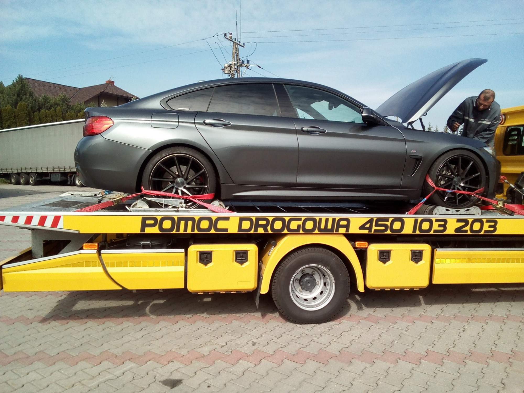 Szary samochód BMW na żółtej lawecie z napisem 'Pomoc Drogowa' w Koziegłowach, mężczyzna w szarej bluzie przy otwartej masce auta.