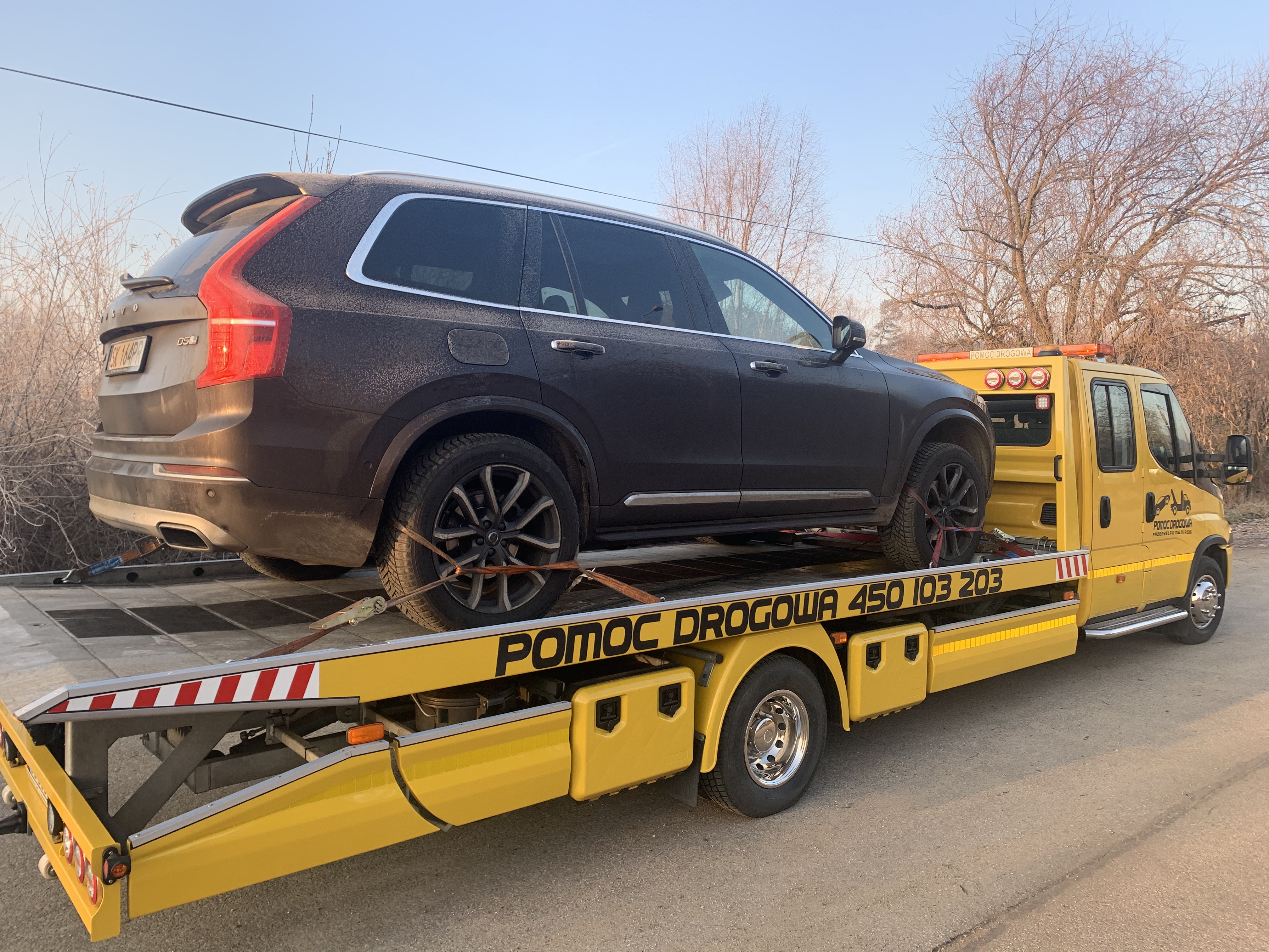 Brązowy SUV Volvo XC90 zabezpieczony pasami na żółtej lawecie z napisem 'POMOC DROGOWA 450 103 203' w słoneczny dzień.