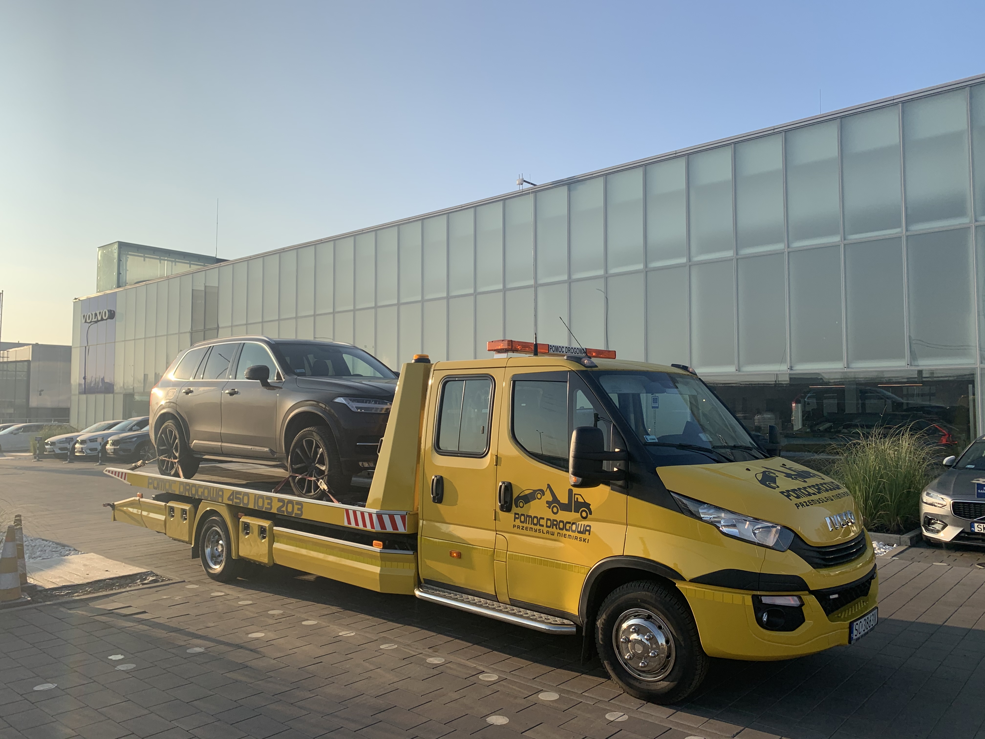Żółta laweta z załadowanym szarym Volvo XC90 na tle szklanego salonu Volvo. Widoczny numer telefonu pomocy drogowej i logo firmy na pojeździe.