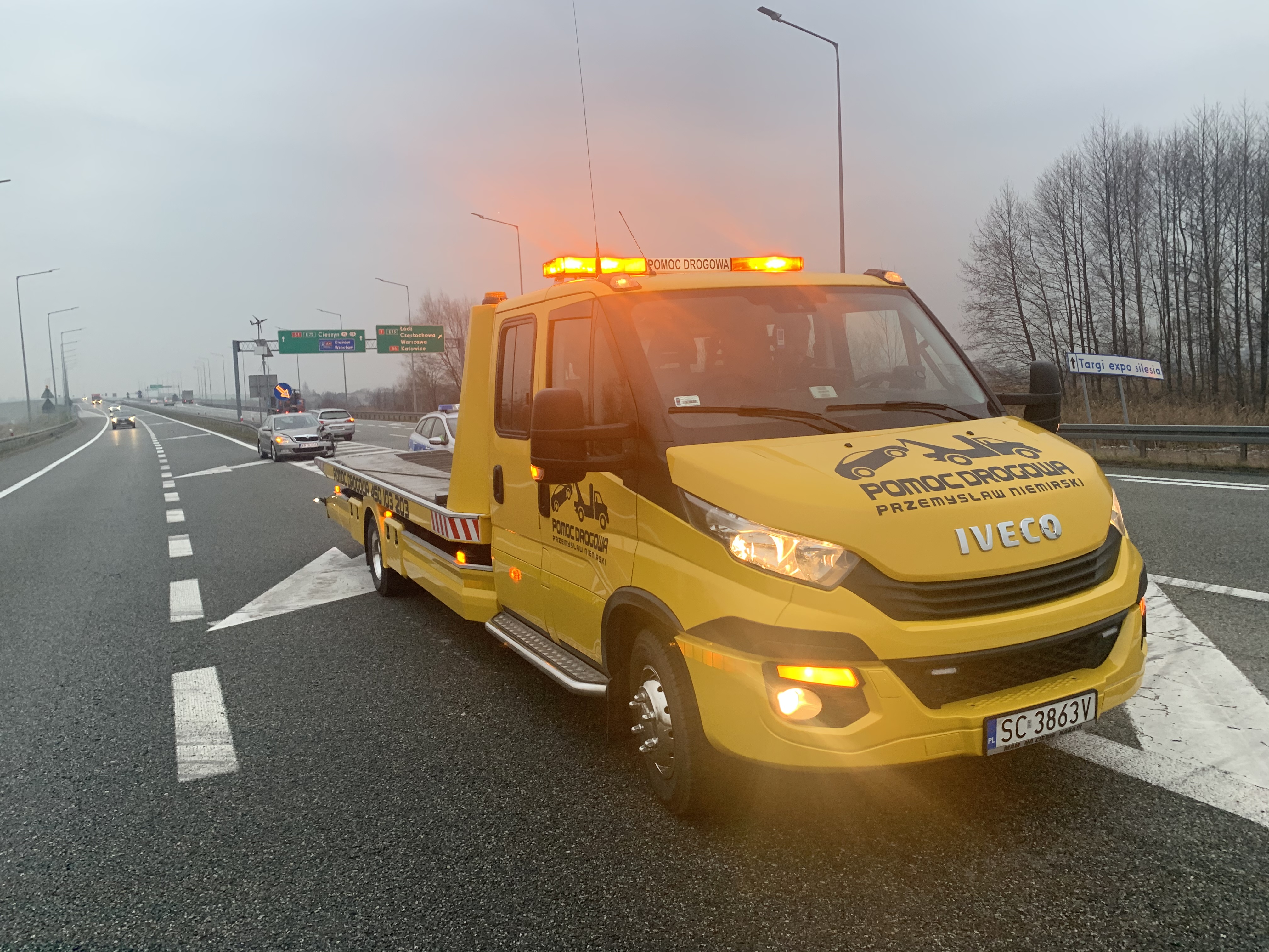 Żółta laweta Iveco z włączonymi światłami ostrzegawczymi na autostradzie, widoczny znak drogowy kierujący na Targi Expo Silesia.
