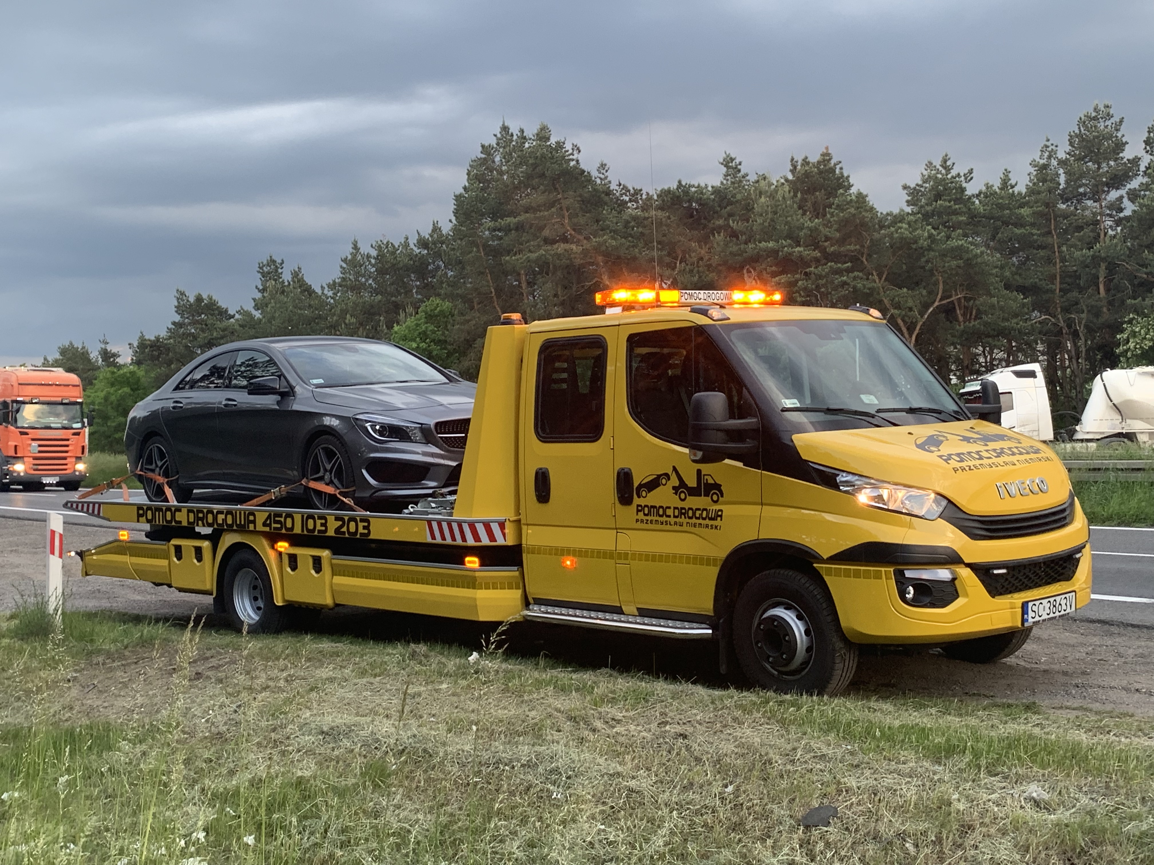 Żółta laweta Iveco z włączonymi światłami ostrzegawczymi przewozi szary samochód osobowy na platformie. W tle widoczny las, pochmurne niebo i inne pojazdy na drodze.