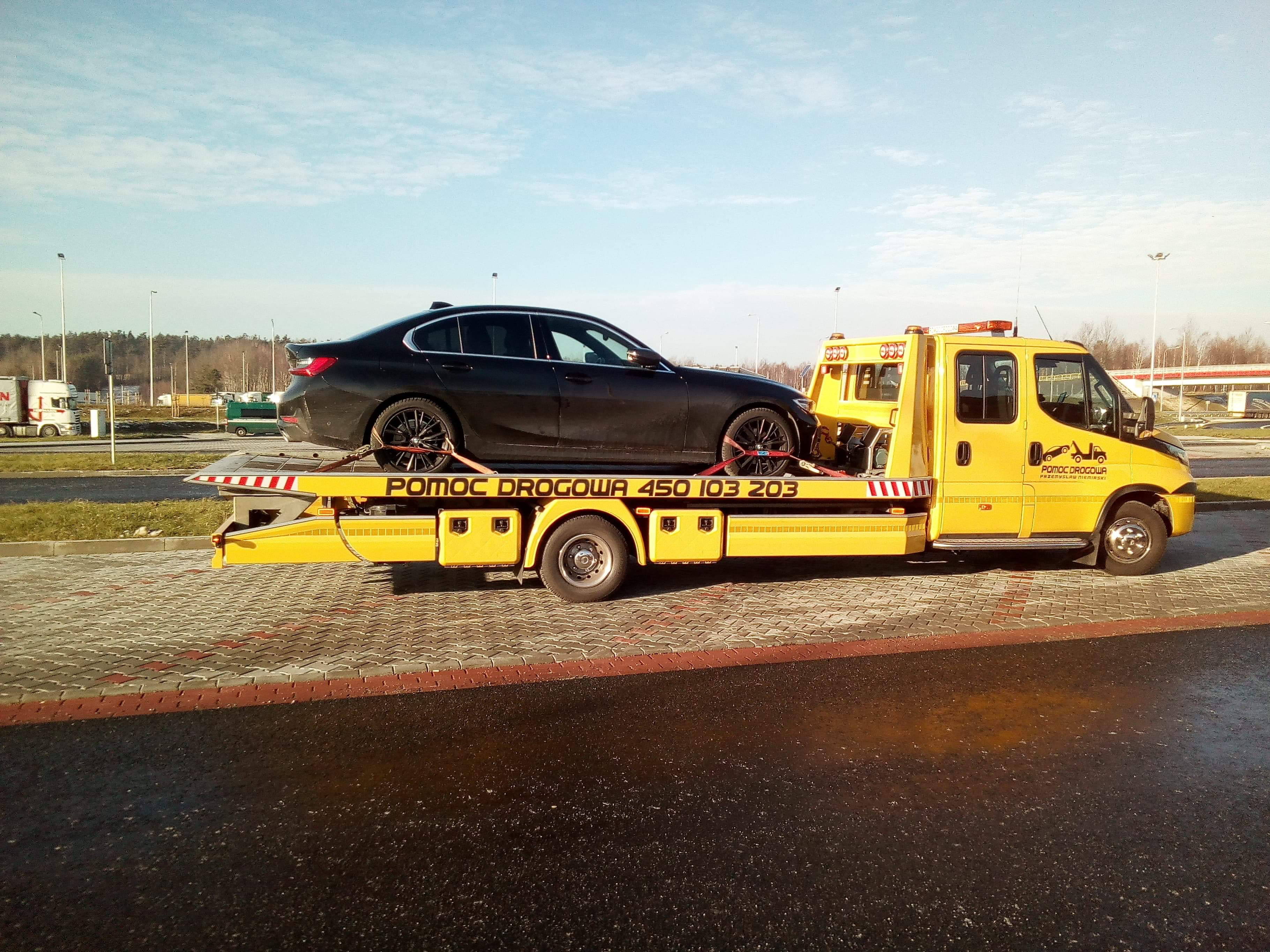 Czarny samochód osobowy marki BMW serii 3 zabezpieczony pasami na żółtej lawecie pomocy drogowej z widocznym numerem telefonu, na parkingu przy autostradzie w słoneczny dzień.