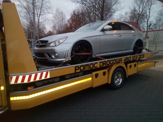 Srebrny Mercedes-Benz CLS na żółtej lawecie z napisem 'POMOC DROGOWA 450 103 203', zabezpieczony czerwonymi pasami transportowymi, widok z boku na tle szarego parkingu i drzew bez liści.