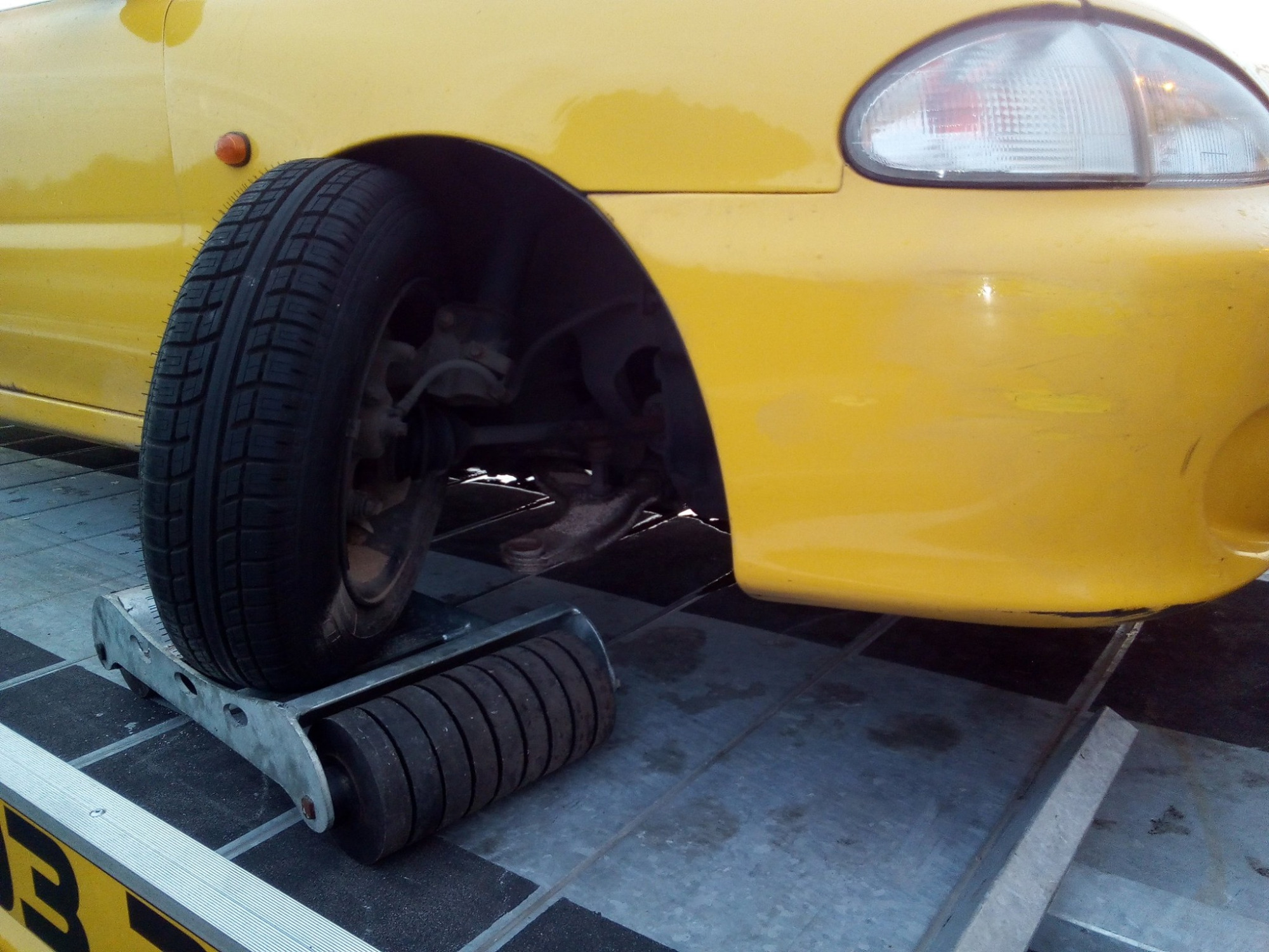 Żółty samochód osobowy, częściowo widoczny, z przednim kołem umieszczonym na rolkach platformy najazdowej, co sugeruje przygotowanie do transportu.