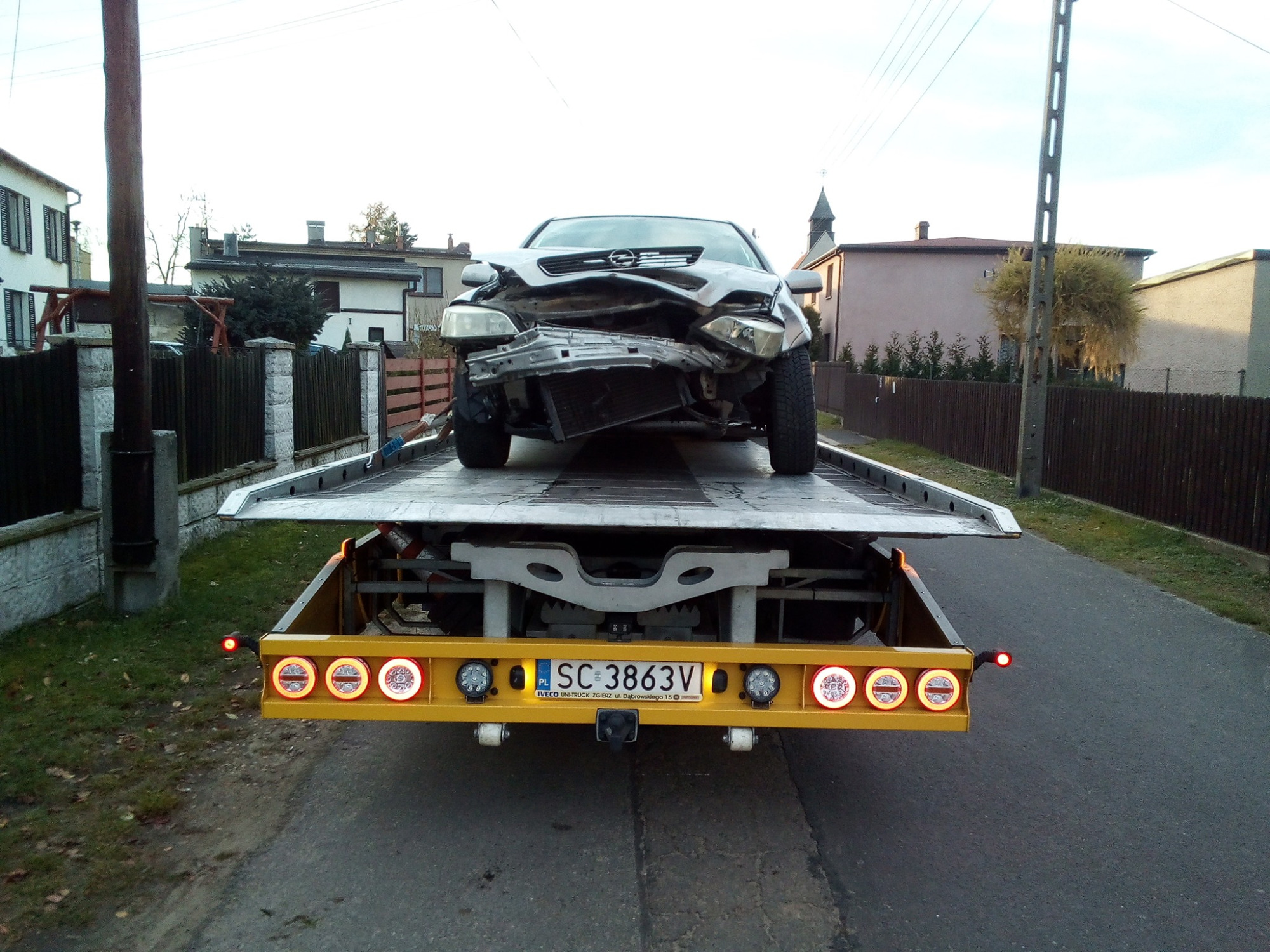 Uszkodzony, srebrny Opel na żółtej lawecie z włączonymi światłami, widok z tyłu na tle wiejskich zabudowań.