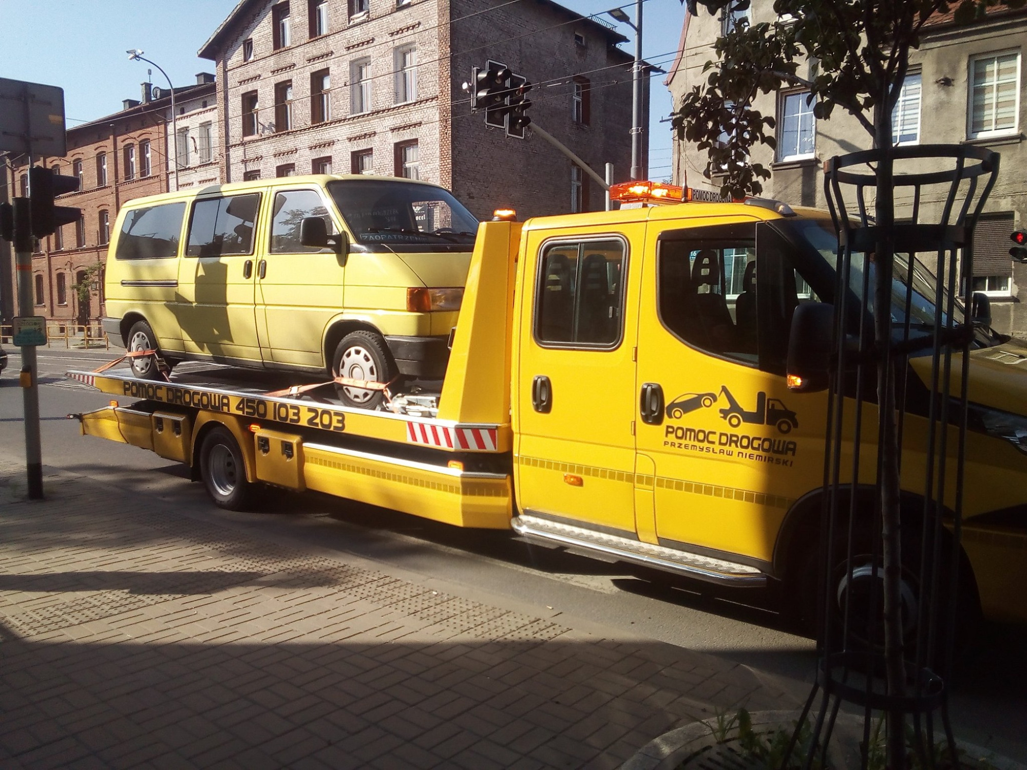 Żółta laweta z załadowanym żółtym busem na tle miejskiej ulicy z sygnalizacją świetlną i budynkami z czerwonej cegły.