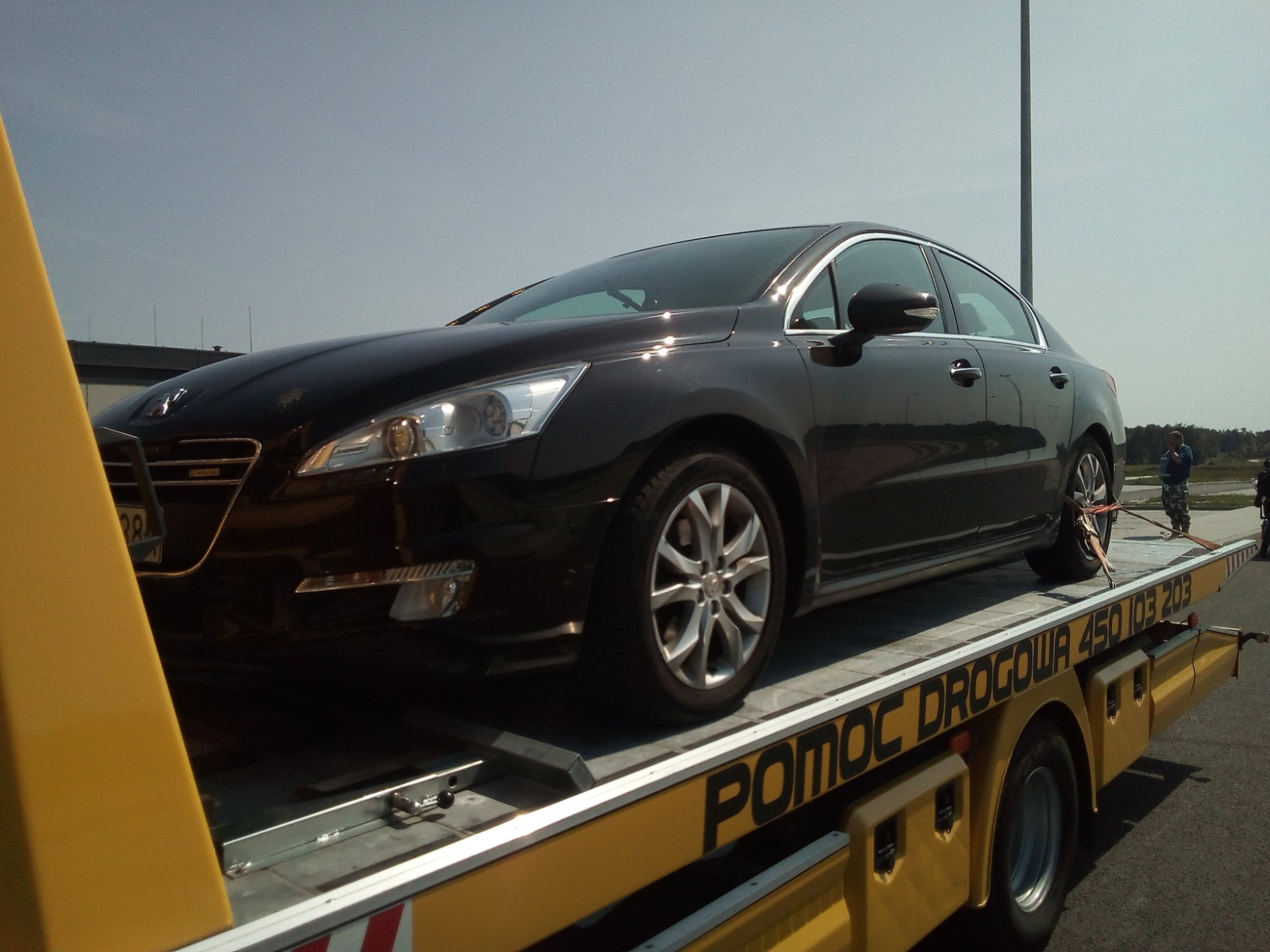 Czarny samochód osobowy marki Peugeot 508 zabezpieczony pasami na żółtej platformie pojazdu pomocy drogowej z napisem 'POMOC DROGOWA', widok z boku w słoneczny dzień.