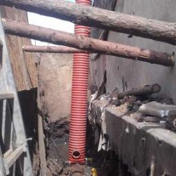 Mm Instal - Pomarańczowa, karbowana rura pionowa, podłączona do brązowej rury poziomej, widoczna w wykopie ziemnym z elementami starej konstrukcji i drewnianymi belkami podtrzymującymi.