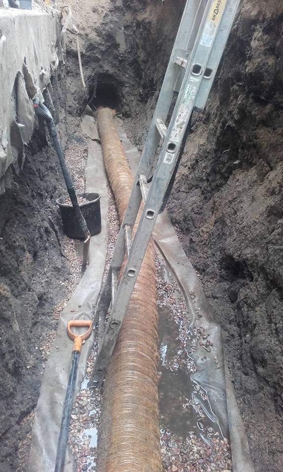 Wykop z ułożoną rurą kanalizacyjną na geowłókninie, obsypaną żwirem, z widoczną drabiną opartą o ścianę wykopu i narzędziami pracy.