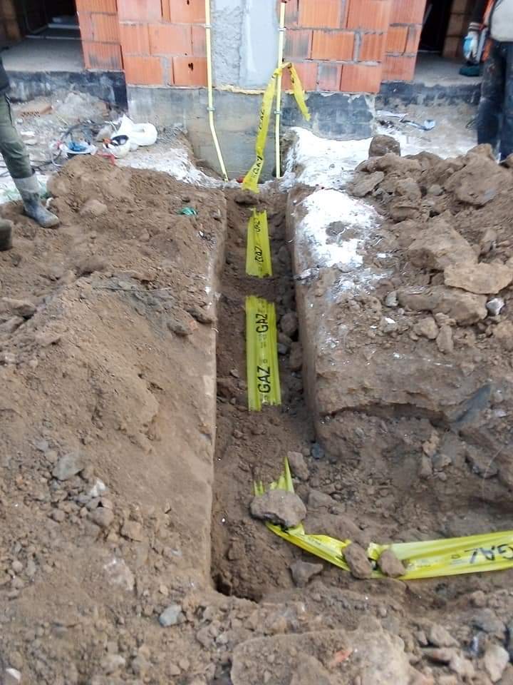 Wykop z żółtą taśmą ostrzegawczą i rurami gazowymi w ziemi, widoczne fundamenty budynku w tle.