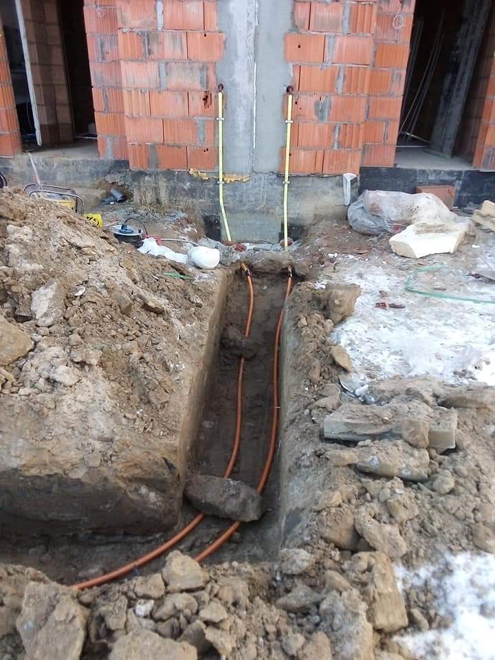 Wykop z pomarańczowymi rurami w ziemi, prowadzącymi do budynku w trakcie budowy z widocznymi żółtymi rurami na ścianie, częściowo zasypany ziemią i kamieniami.