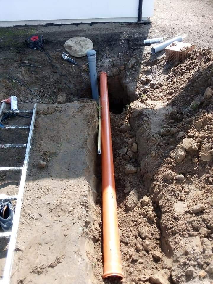 Wykop z rurą kanalizacyjną w ziemi, widoczne narzędzia i cegły, instalacja przy budynku.