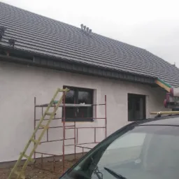 Nowy dach z szarej dachówki ceramicznej, widoczne kominki wentylacyjne, obróbki blacharskie i częściowo otynkowana ściana budynku z oknami, rusztowanie i drabina przy elewacji, widok zza szyby...