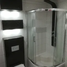 Nowoczesna łazienka z kabiną prysznicową narożną z czarnym panelem natryskowym, podwieszaną toaletą i czarnymi szafkami podświetlanymi od dołu, wykończona białymi płytkami.