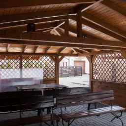 Drewniana altana z ażurowymi ścianami, wyposażona w stół i ławki, widok na posesję z kostką brukową i ogrodzeniem.