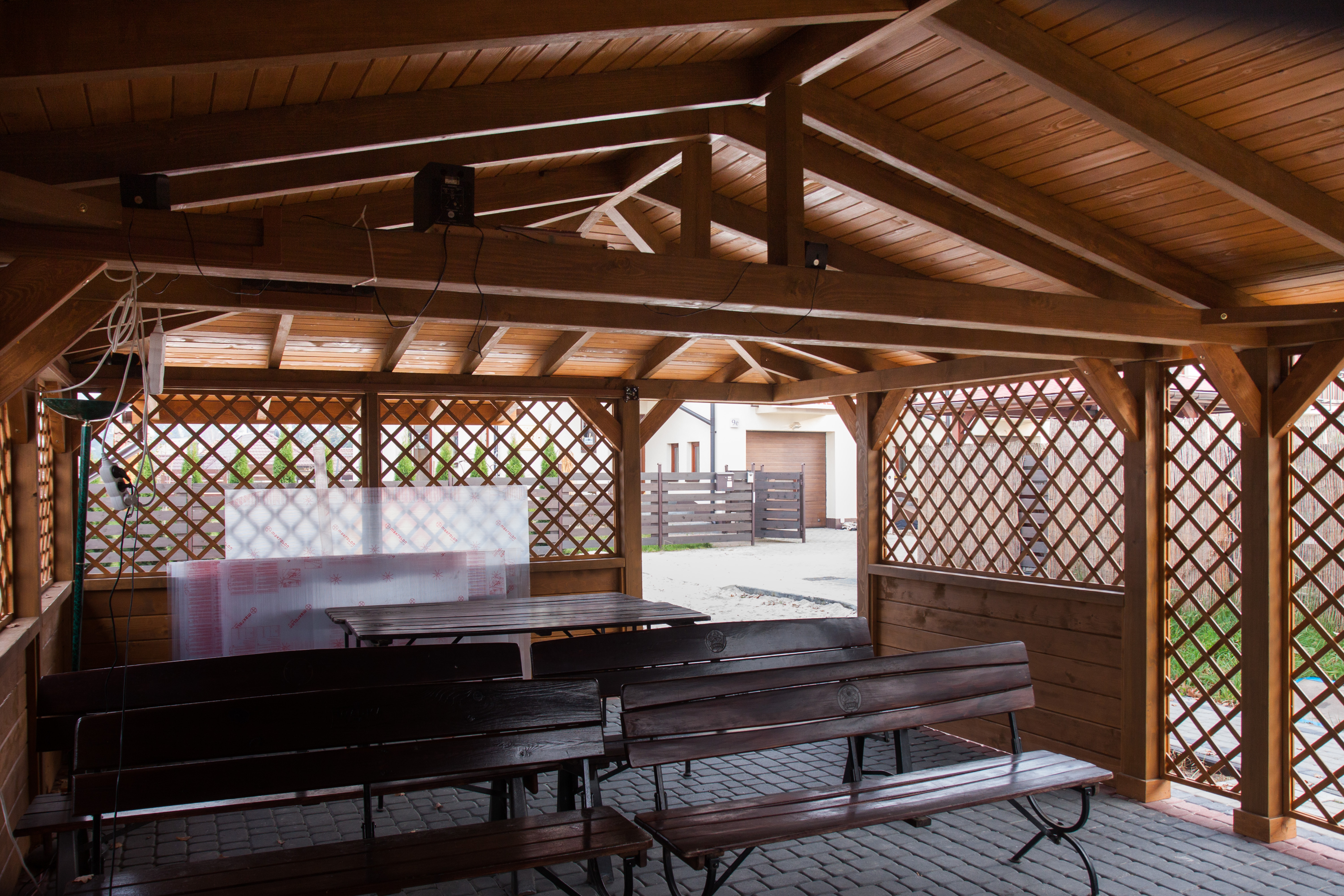 Drewniana altana z ażurowymi ścianami, wyposażona w stół i ławki, widok na posesję z kostką brukową i ogrodzeniem.