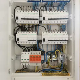 Otwarta skrzynka elektryczna z zainstalowanymi bezpiecznikami, wyłącznikami różnicowoprądowymi i listwami zaciskowymi, widoczne kable w różnych kolorach, zamontowane w białej obudowie.