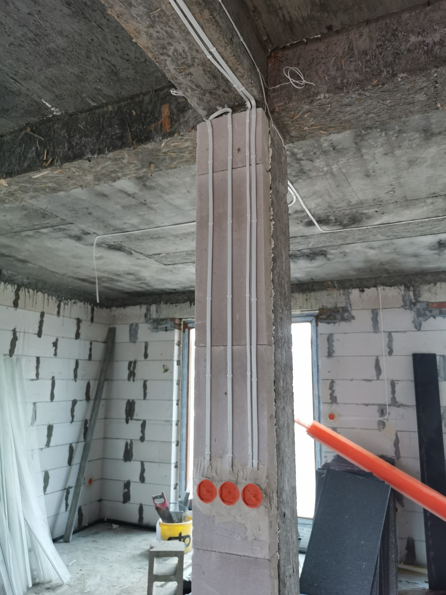 Wewnętrzna instalacja elektryczna w surowym stanie: białe przewody prowadzone wzdłuż betonowego słupa, widoczne puszki elektryczne, surowy strop i ściany z betonu komórkowego.