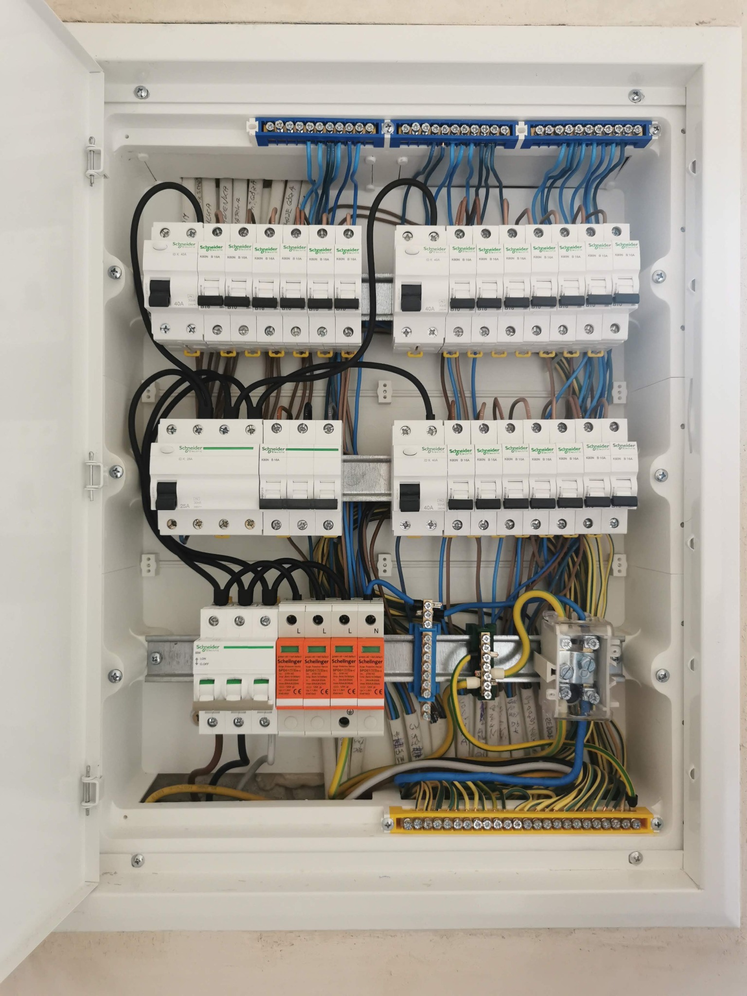 Otwarta skrzynka elektryczna z zainstalowanymi bezpiecznikami, wyłącznikami różnicowoprądowymi i listwami zaciskowymi, widoczne kable w różnych kolorach, zamontowane w białej obudowie.