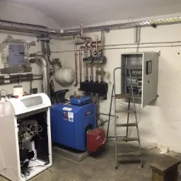 Piwnica z dwoma kotłami grzewczymi, skrzynką elektryczną na ścianie, instalacją rur miedzianych pod sufitem i drabiną. Jeden z kotłów jest otwarty, ukazując wewnętrzne komponenty. W tle widoczna...