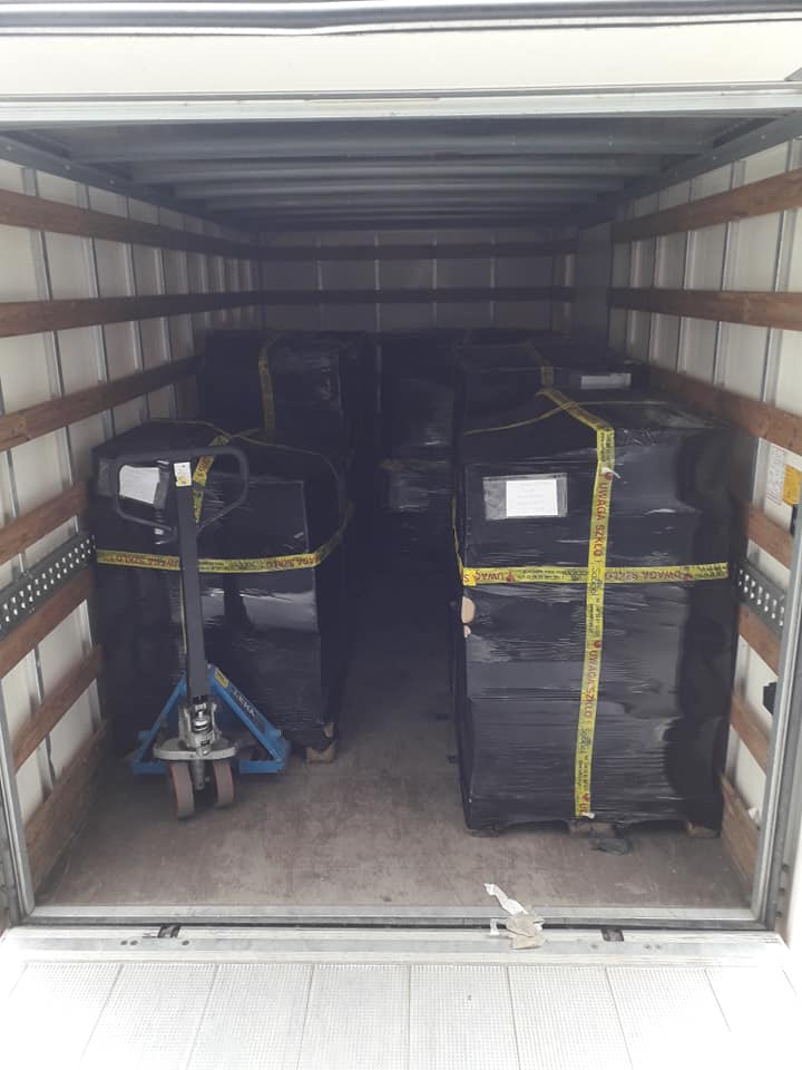 Wnętrze przestrzeni ładunkowej z paletami owiniętymi czarną folią i zabezpieczonymi żółtą taśmą ostrzegawczą, w tle widoczny wózek paletowy.