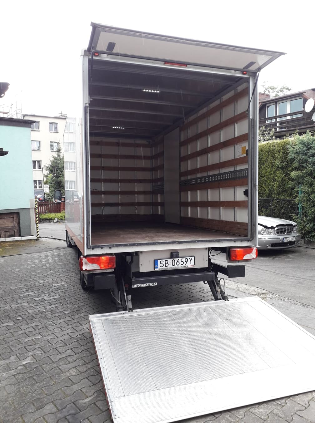 Otwarty, pusty samochód dostawczy z podniesioną rampą załadunkową na kostce brukowej, widok z tyłu na tle budynków mieszkalnych i zaparkowanego samochodu osobowego.
