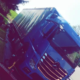 Niebieski samoch&oacute;d ciężarowy Mercedes-Benz Actros z naczepą, widziany z przodu pod kątem, z widoczną tablicą rejestracyjną KR 6FV67, zaparkowany na poboczu z trawą i drzewami w tle.