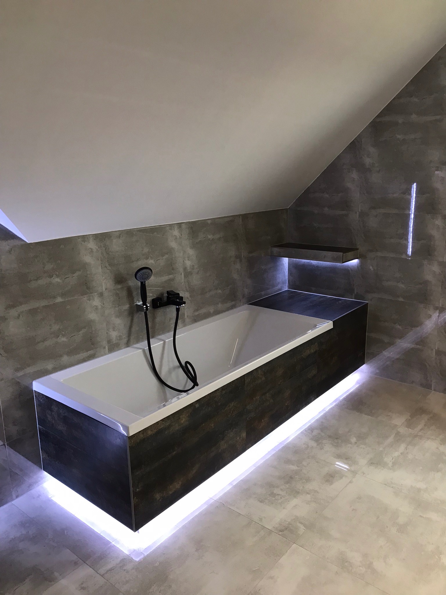 Nowoczesna łazienka na poddaszu z wanną obudowaną płytkami imitującymi drewno, podświetloną listwą LED, szare płytki na ścianach i podłodze, czarna armatura i półka z podświetleniem.