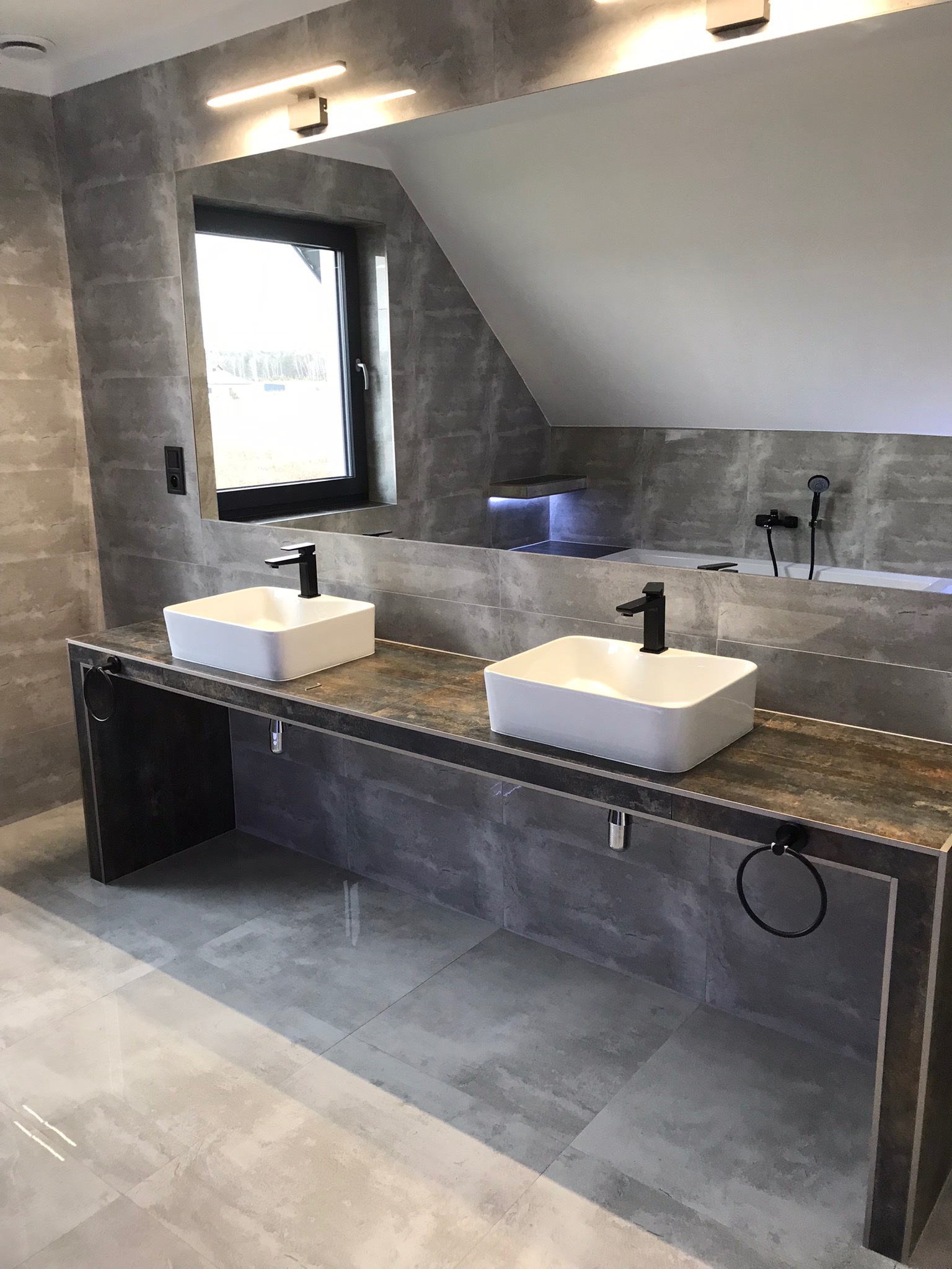 Nowoczesna łazienka z dwoma kwadratowymi umywalkami nablatowymi, czarną armaturą i szarą płytką imitującą beton, z oknem w tle i dużym lustrem.