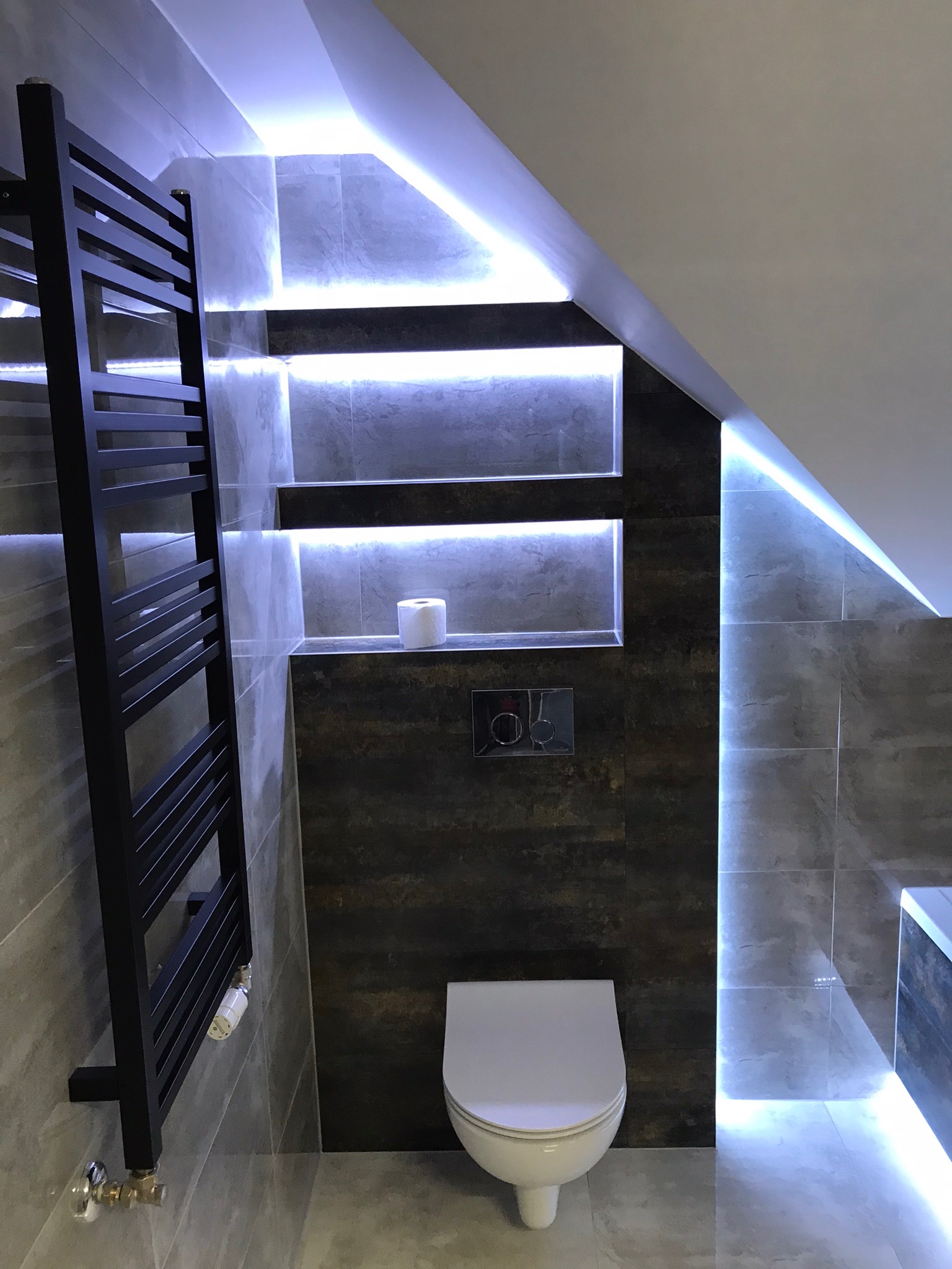 Nowoczesna łazienka w szarościach z podświetlanymi półkami wnękowymi, czarnym grzejnikiem drabinkowym i podwieszaną toaletą.