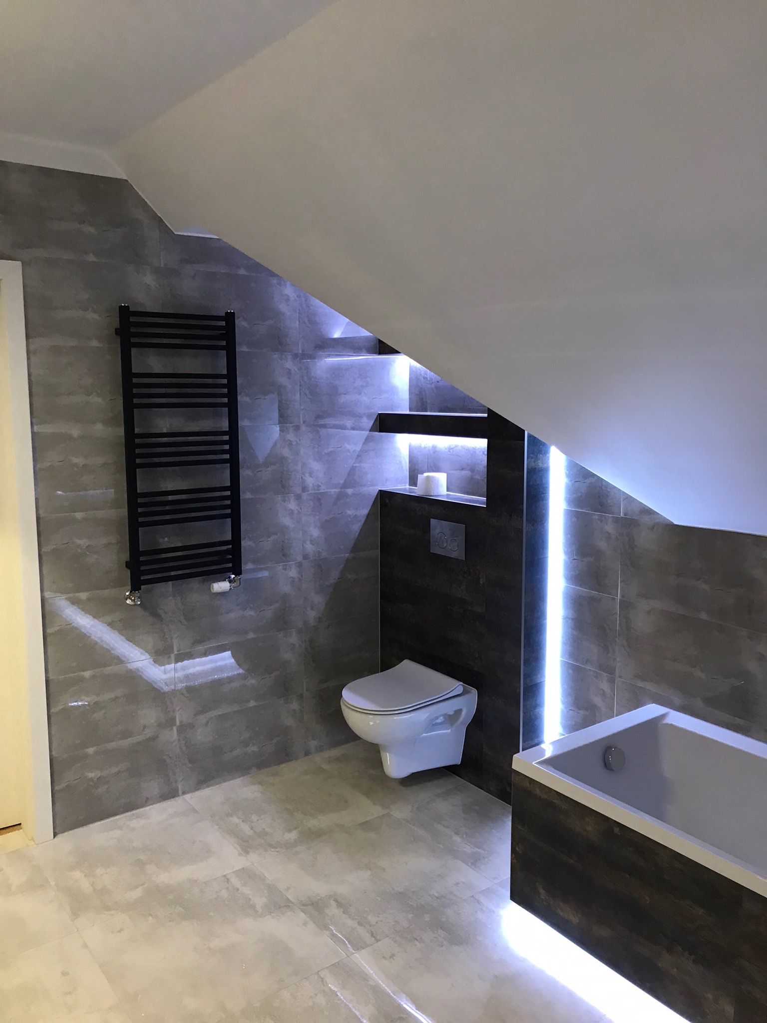 Nowoczesna łazienka na poddaszu z szarymi płytkami, podświetlaną wnęką i wanną z drewnopodobną obudową oraz czarnym grzejnikiem drabinkowym.