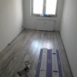 Wnętrze pokoju w trakcie układania paneli podłogowych z widocznymi kartonami paneli i elementami dystansowymi przy ścianie