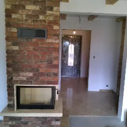 Murowany kominek z cegły klinkierowej w nowym domu, widoczny w trakcie wykańczania wnętrza, z drewnianymi belkami stropowymi i błyszczącą podłogą z płytek.