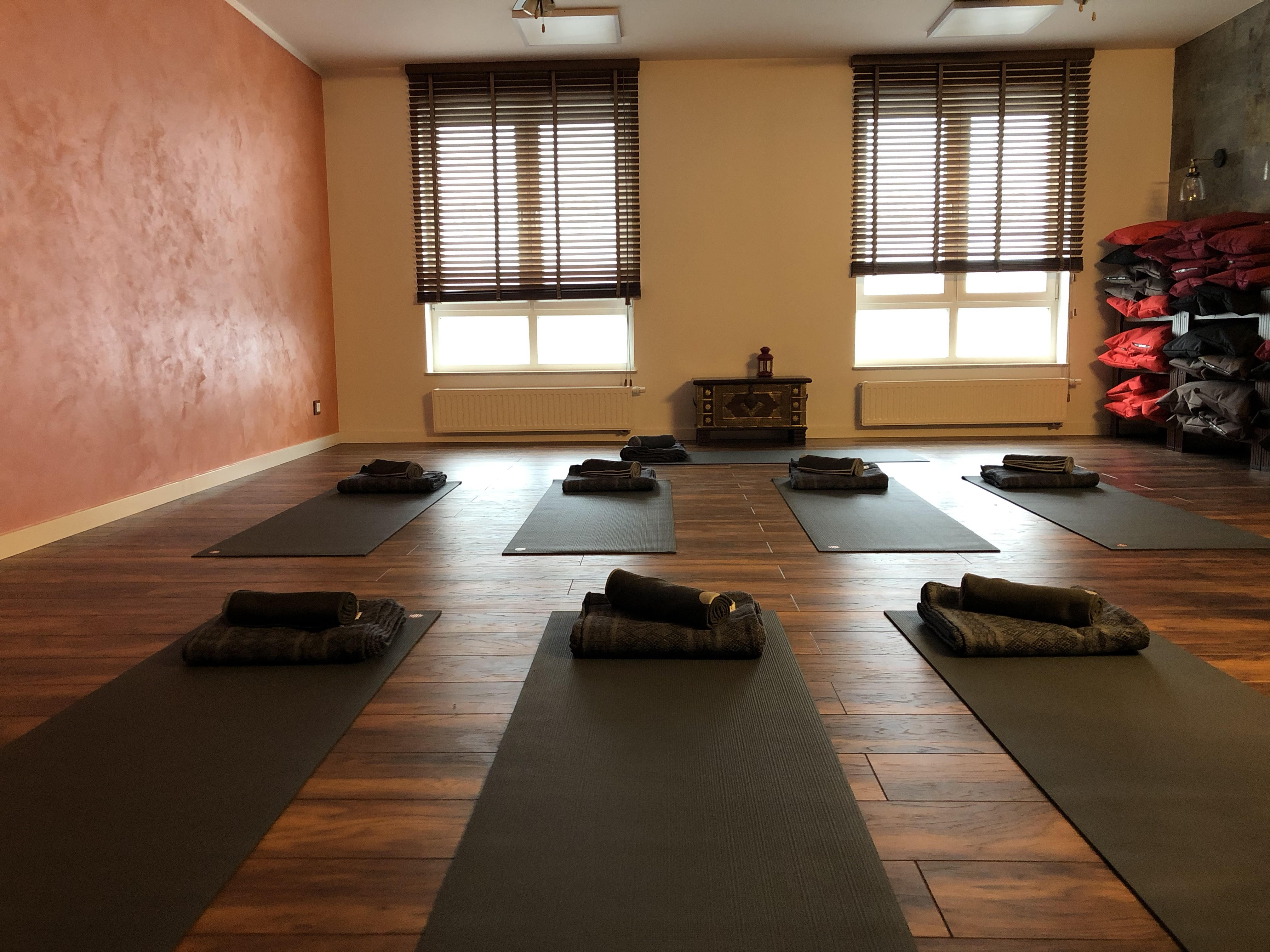 Sala do jogi z rozłożonymi matami, zwiniętymi ręcznikami i poduszkami, drewnianą podłogą i ścianą w kolorze terakoty.