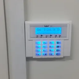 Biały panel alarmowy Safet Versa z podświetlanym niebieskim ekranem LCD i klawiaturą, zamontowany na białej ścianie obok drzwi z zamkiem.