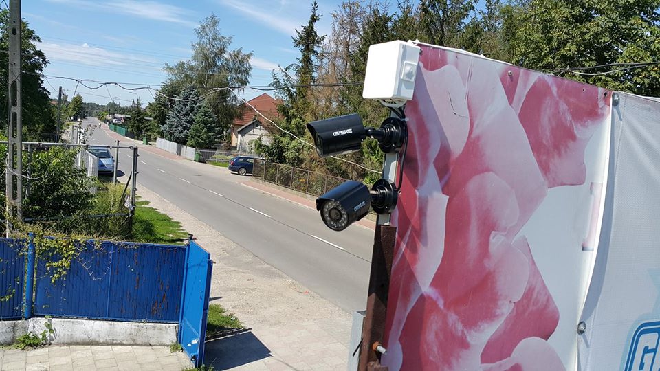 Dwie czarne kamery monitoringu marki GISE zamontowane na konstrukcji z reklamą kwiatową, skierowane w stronę ulicy z widocznymi domami i samochodami w tle.