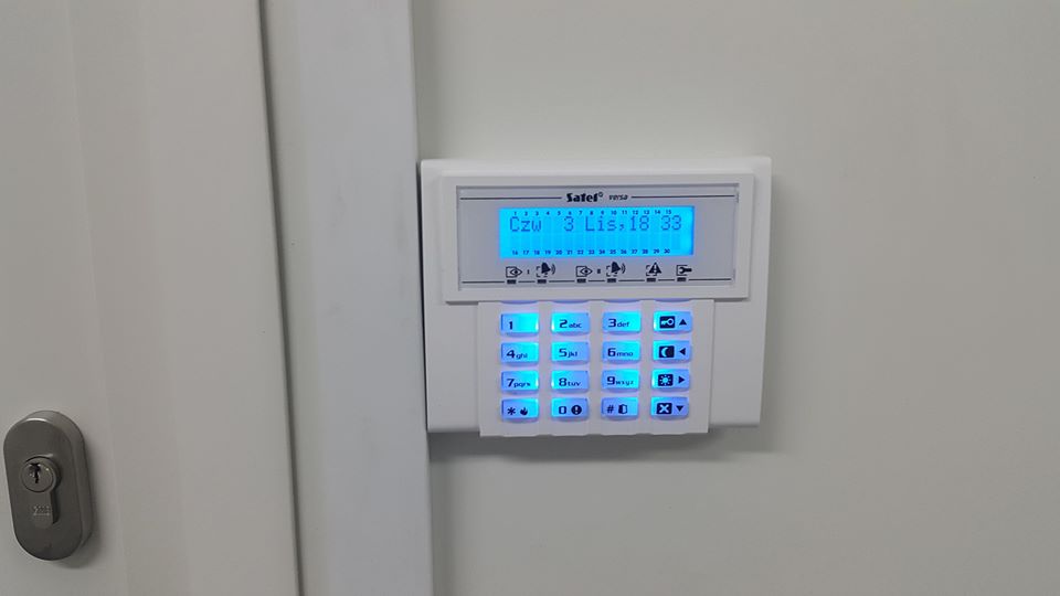 Biały panel alarmowy Safet Versa z podświetlanym niebieskim ekranem LCD i klawiaturą, zamontowany na białej ścianie obok drzwi z zamkiem.