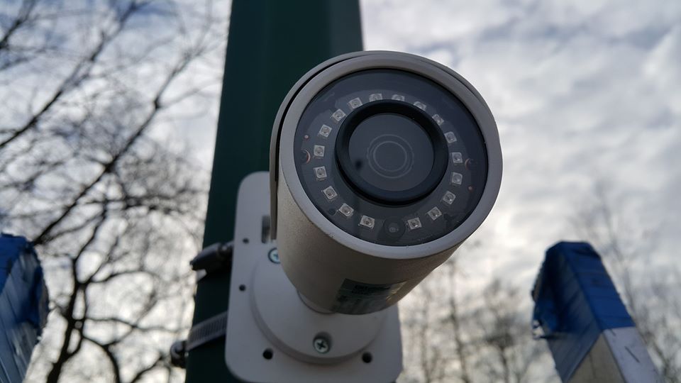 Kamera monitoringu zewnętrznego zamontowana na słupie w Otwocku, widok z dołu na tle pochmurnego nieba i drzew bez liści.