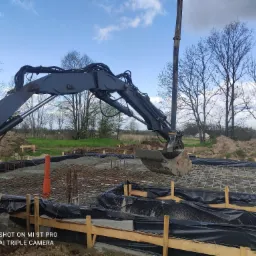 Wykop pod fundamenty w Nysie: Koparka z łyżką nad zbrojeniem i częściowo zalanym betonem, widoczne szalunki i folia izolacyjna.