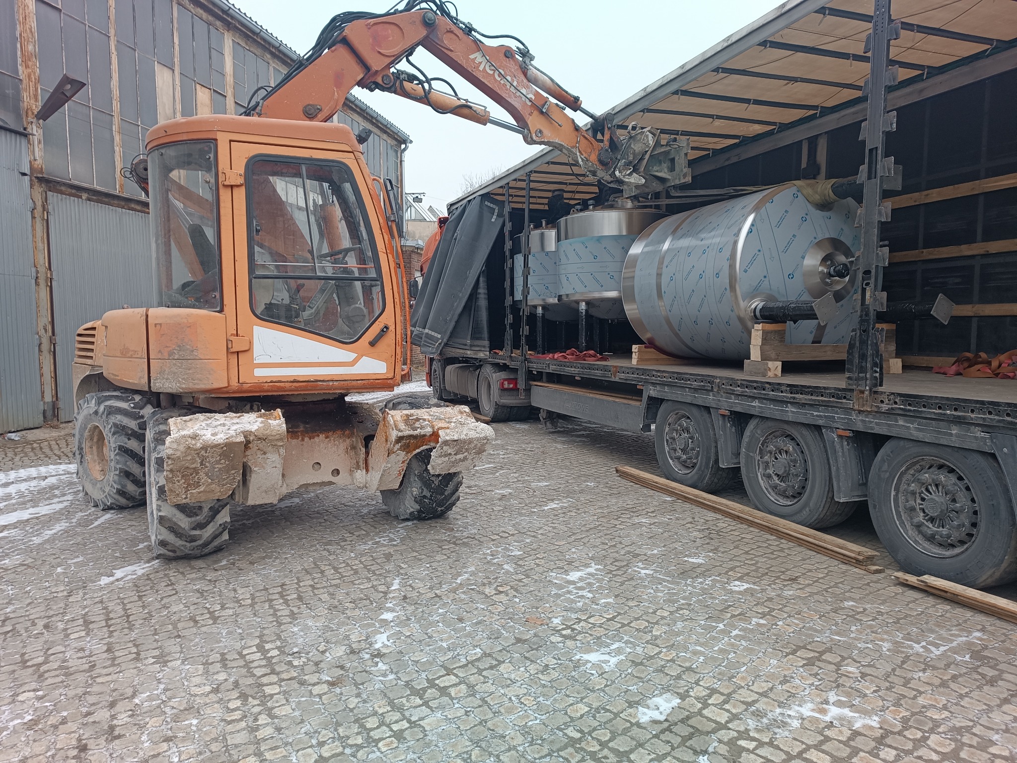 Pomarańczowa koparka kołowa z łyżką podnosi srebrny zbiornik na naczepę ciężarówki na tle budynku przemysłowego i kostki brukowej.