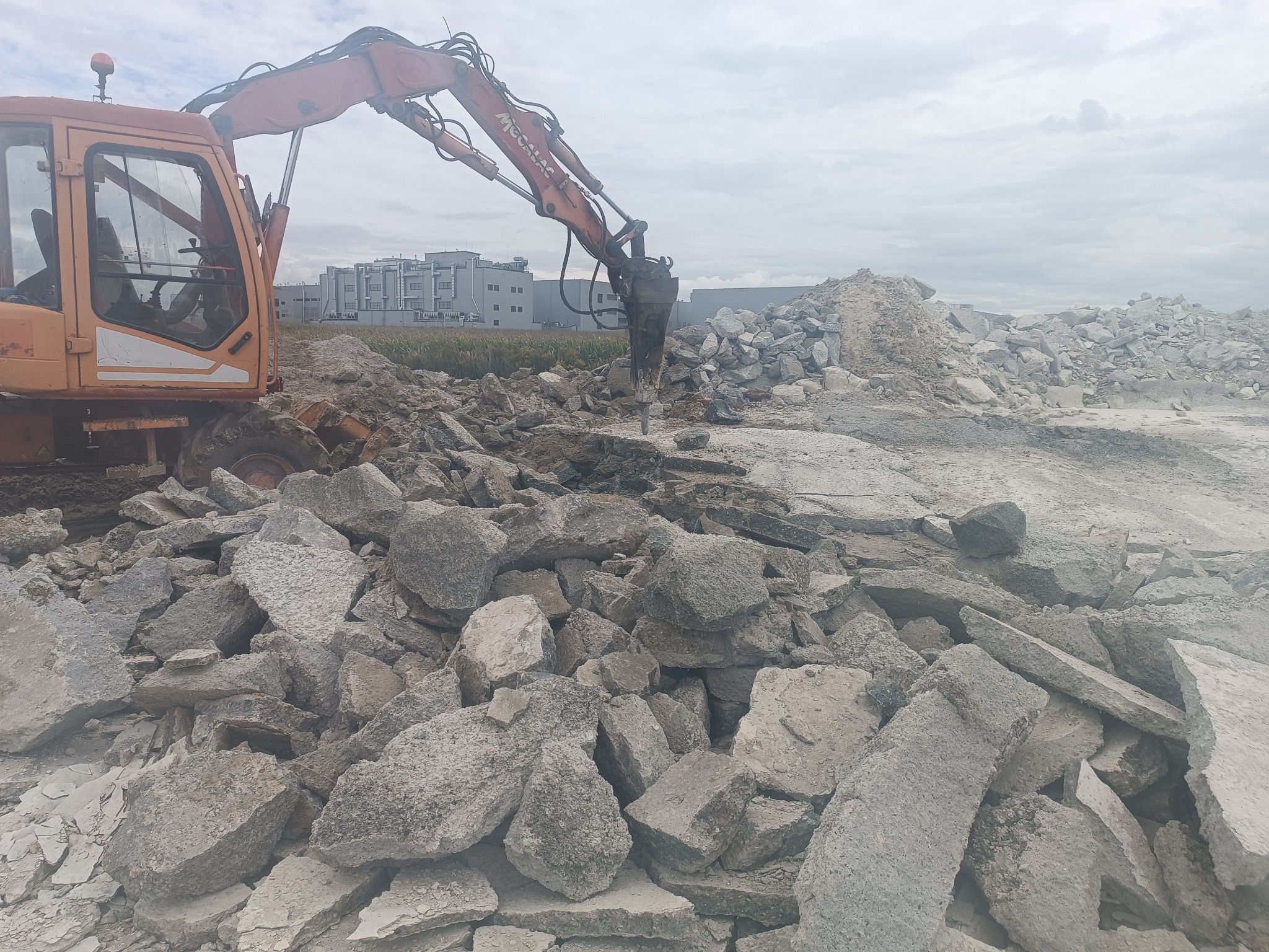 Pomarańczowy koparko-ładowarka z młotem hydraulicznym kruszy betonowe płyty na placu budowy. W tle budynek i pole uprawne pod pochmurnym niebem.