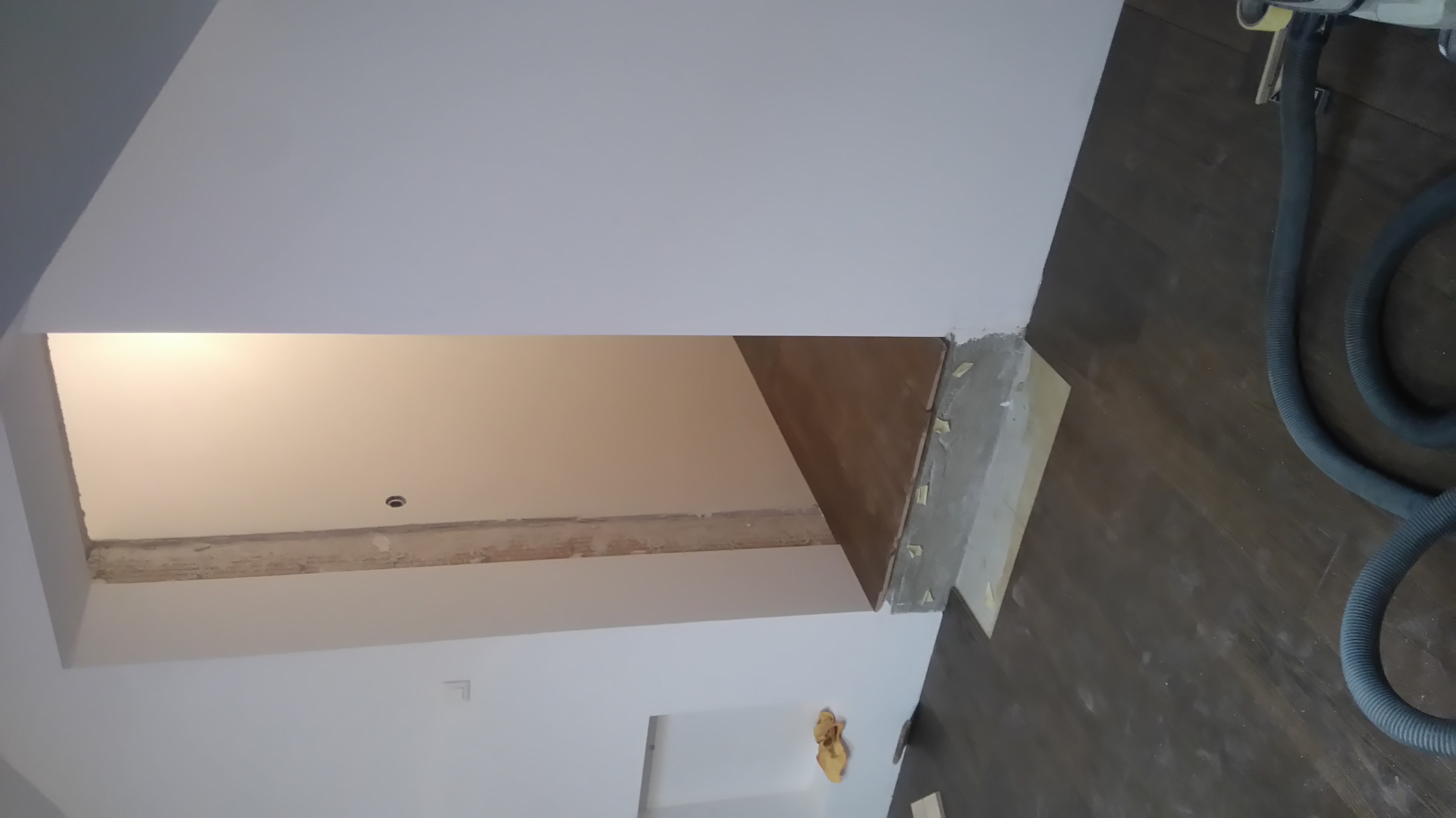 Fragment pokoju z ciemną drewnianą podłogą w trakcie wykańczania, widoczne narzędzie do szlifowania z podłączonym odkurzaczem, ściany białe, w tle widoczny fragment innego pomieszczenia.