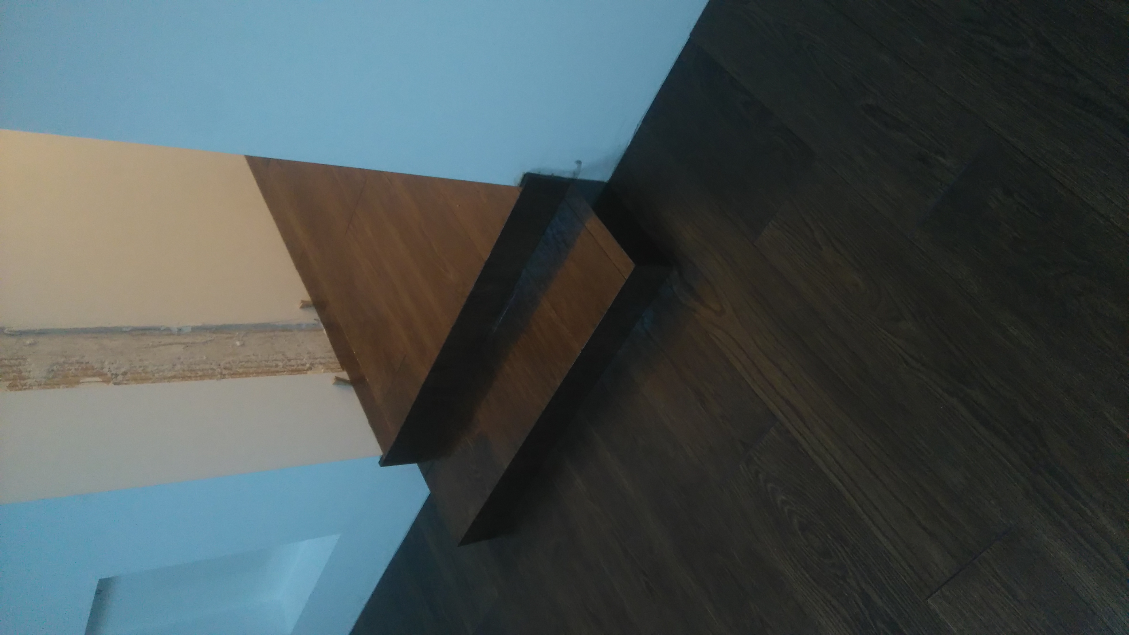 Fragment ściany z widocznym śladem po starej listwie przypodłogowej oraz nowa, drewniana listwa w kolorze podłogi z ciemnych paneli. Podłoga ułożona wzdłuż ściany.