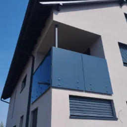 Nowoczesny budynek z balkonem zabezpieczonym szklaną balustradą w odcieniach szarości, widok z dołu na błękitne niebo.