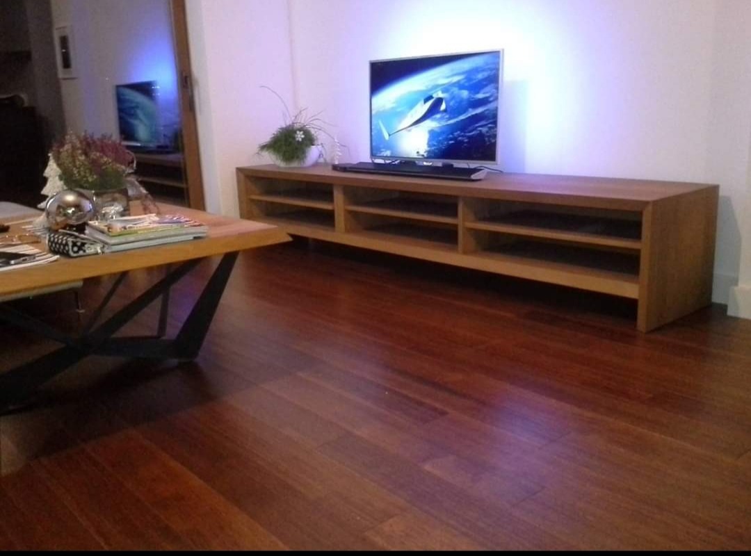 Drewniana podłoga z egzotycznego drewna prowadzi do niskiej szafki RTV z otwartymi półkami, na której stoi telewizor i roślina doniczkowa. W tle widoczne lustro.