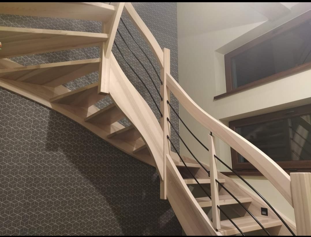 Spiralne schody z jasnego drewna z czarnymi, metalowymi balustradami na tle ściany z geometrycznym wzorem i dwoma oknami.