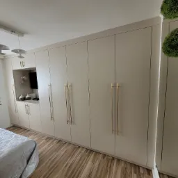 Beżowa szafa wnękowa z długimi, złotymi uchwytami w sypialni, widok z boku łóżka, na podłodze panele imitujące drewno.